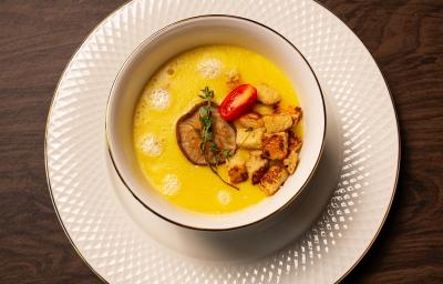 Fotografia em tons de marrom com um prato de sopa branco ao centro. Dentro do prato existe um creme de cor amarela feito com mandioquinhas acompanhado com croutons, cogumelos e tomate cereja.