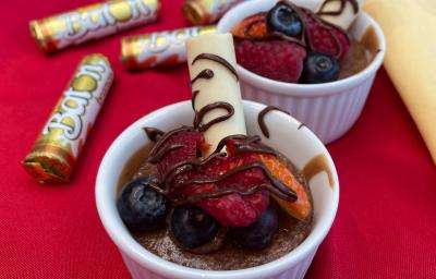 Fotografia em tons de vermelho com dois refratários ao centro. Dentro de cada um deles existe um bolo de chocolate cremoso coberto com morangos, calda de chocolate e BATON branco. Ao fundo existe embalagens de BATON branco.