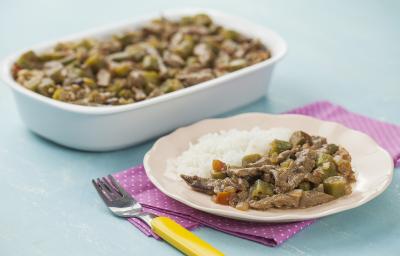 Fotografia em tons de azul e rosa em uma bancada de madeira azul, um guardanapo de pano rosa com bolinhas brancas, um refratário branco retangular com o picadinho de carne com quiabo. Ao lado, um prato rosa redondo com uma porção do picadinho e arroz.