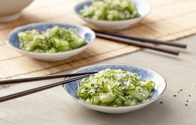 Fotografia em tons de verde em uma bancada de madeira com uma toalha de palha bege. Ao centro, um prato japonês branco com detalhes em azul e a salada de sunomono (pepinos fatiados bem finos) dentro, com um par de hashi apoiado.