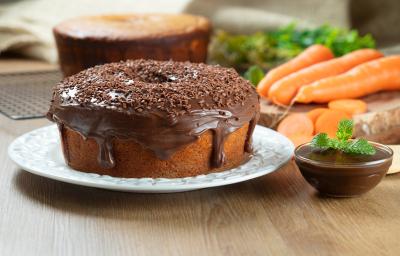 Fotografia de dois bolos de cenoura, um sem e o outro com cobertura de chocolate cremoso e granulado, apoiado em um prato branco. Ao lado tem mais cobertura em um recipiente pequeno de vidro, e ao fundo tem cenouras inteiras e alguns pedaços.