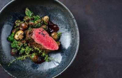 Fotografia vista de cima de um de assado ao molho ferrugem delicioso feito com Filé mignon temperado, com molho de vinho tinto e champignons. A receita está em um prato fundo de tom azul escuro, sobre uma mesa de cimento queimado escuro.