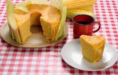 Foto de uma mesa com toalha vermelha quadriculada com dois pratos redondos, um com um bolo de fubá sem uma fatia, decorado com as folhas da espiga, e outro com uma fatia cortada em frente a uma xícara de café de louça vermelha