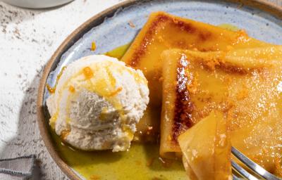 Foto de um prato em tom claro com a receita de Crepe Suzette, servido com calda amarela e uma bola de sorvete. O prato está em uma bancada clara, com. um pote de calda amarela e um guardanapo ao lado.