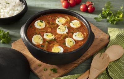 Fotografia em tons de verde em uma bancada de madeira verde, uma tábua de madeira, um paninho verde com bolinhas brancas, uma panela de barro grande ao centro com a moqueca de ovos dentro dela. Ao fundo, um recipiente com arroz, tomates e salada verde.