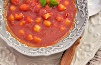 Fotografia vista de cima de uma sopa de tomates com nhoquettes em um prato fundo desenhado em tons de cinza, apoiado sobre um prato raso branco desenhado com uma colher apoiada. Os pratos estão sobre uma toalha de mesa marrom.