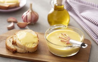 Foto em tons de amarelo da receita de aioli servida em um refratário de vidro redondo com um fuet mergulhado e fatias de pão ao lado com o o aioli, tudo sobre uma tábua de madeira. Ao fundo há um vidro de azeite e uma cabeça de alho