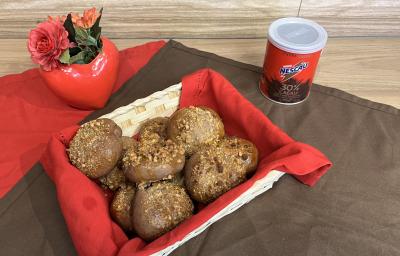 Fotografia de pãezinhos de nescau com castanhas dentro de uma cestinha branca quadrada com um pano vermelho. A cestinha está sobre um pano marrom de tom escuro, ao lado da latia de nescau extra cacau.