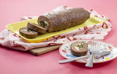 Fotografia em tons de rosa em uma mesa rosa com um pano rosa desenhado e um recipiente retangular amarelo com o bolo de carne com espinafre em cima dele. Ao lado, um prato colorido com uma fatia do bolo e um garfo e uma faca apoiados sobre ele.