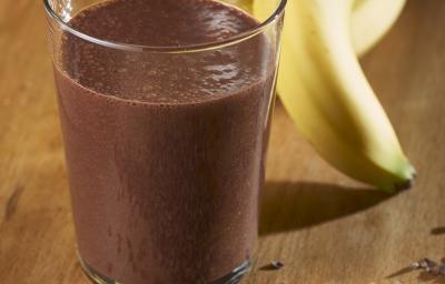 Fotografia de um copo de vidro com uma vitamina de banana e chocolate Nescau. Ao fundo, um cacho de bananas, e na frente do copo, raspas de chocolate sobre uma mesa de madeira.