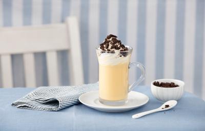 fotografia em tons de azul e branco de uma bancada azul vista de frente, contém um pratinho redondo branco com um copo transparente com a bebida dentro e por cima chantilly e biscoitos triturados para decorar. Ao lado um pano azul e do outro uma colher.