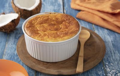 Foto da Receita de Suflê de Coco. Observa-se um recipiente redondo com o suflê sobre uma tábua de madeira redonda. Ao lado do potinho, uma colherinha também de madeira. Ao fundo, um coco aberto e um guardanapo laranja ambientam a foto.