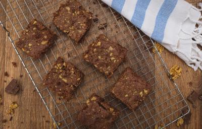 Fotografia em tons de azul em uma bancada de madeira escura, um suporte de grelha prata com alguns brownies de chocolate com nozes em cima dele e ao lado um paninho branco com listras azuis.