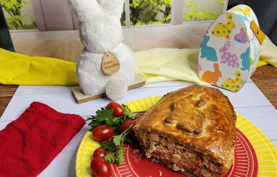 Foto da receita de Ovo de Colher Torta de Frango, servido cortado em um prato vermelho, sobre outro prato amarelo, em uma bancada branca decorada com itens de páscoa, como um coelho branco e uma caixinha em formato de ovo de páscoa