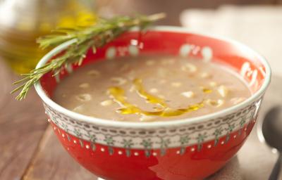 Fotografia em tons de vermelho em uma mesa de madeira com um recipiente redondo e fundo vermelho com a sopa de feijão dentro. Ao lado, um guardanapo de pano bege com uma colher apoiada nele e ao fundo, um potinho de azeite.