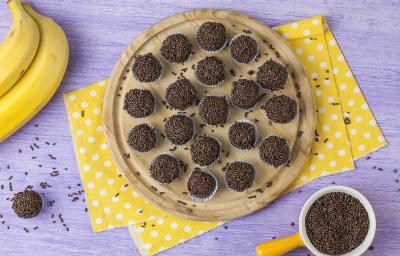 Fotografia em tons de marrom, amarelo e lilás, com bancada lilás vista de cima, ao centro peça de madeira com brigadeiros sobre guardanapo amarelo com bolinhas brancas, ao lado potinho com granulado, um brigadeiro e e duas bananas.