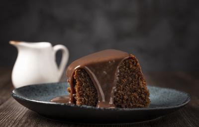 fotografia em tons de preto e marrom tirada de uma fatia de bolo de chocolate com calda por cima, e ao fundo um jarra branca