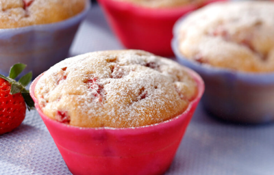 Fotografia em tons de vermelho e lilás em uma bancada com uma toalha emborrachada lilás, com forminhas pequenas vermelhas e lilás com os muffins de fubá com morango dentro delas.