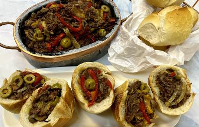 Foto da receita de carne louca servida em uma panela de pedra circular com 5 lanches de carne louca feitos a frente com pão francês e a receita dentro. Ao lado ainda há um saco de pães franceses