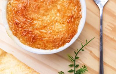 Foto da receita de Cocada de Forno. Observa-se um recipiente redondo branco com a cocada dentro assada e bem douradinha, sobre uma tábua de madeira