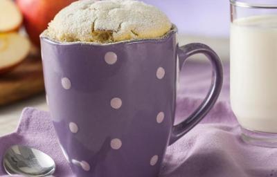 Fotografia em tons de roxo em uma bancada de madeira e um paninho roxo. Ao centro, uma caneca roxa de bolinhas brancas com o bolinho de caneca de leite Ninho em Pó. Ao fundo, maçãs cortadas ao meio e um copo de vidro com leite.