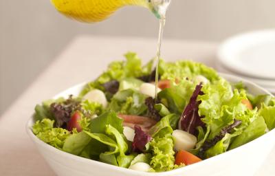 Fotografia em tons de verde e branco de uma bancada de madeira branca com um  recipiente branco quadrado com salada de folhas e um fio de azeite sendo despejado. Ao lado uma tabua de madeira com uma colher de servir.