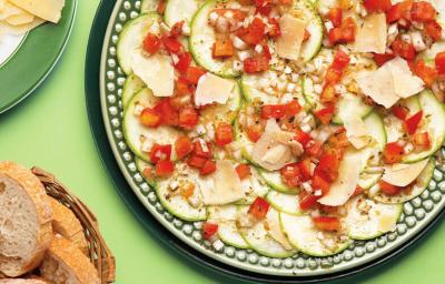 Fotografia em tons de verde e vermelho em uma mesa verde e um prato ao centro com o carpaccio de abobrinha. Ao lado, uma cesta de vime com fatias de pães e um prato verde com lascas de queijo parmesão.