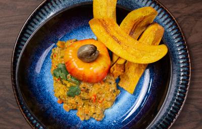 Fotografia em tons de marrom com um prato preto ao centro. Em cima do prato existe uma receita de ceviche de caju com castanhas e chips de banana da terra