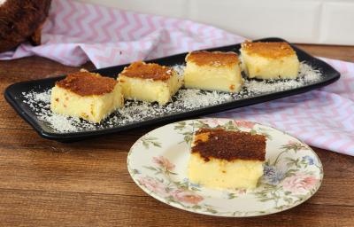 Foto da receita de Docinho do Céu Cremoso. Observa-se um recipiente preto com quatro retângulos do doce polvilhado de coco e, à frente, um pratinho de sobremesa com uma fatia também.