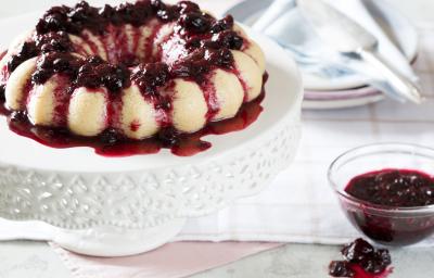 foto em tons de branco e vinho tirada de uma mesa branca, contém um suporte para servir sobremesas branco com o Brigadeirão Branco com Calda de Frutas Vermelhas. Ao lado um pote transparente redondo com a calda de frutas vermelhas.