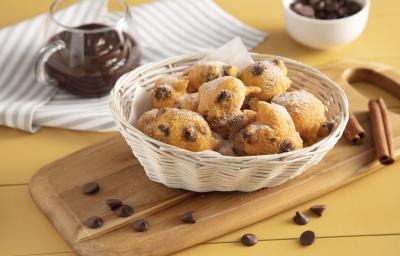 Fotografia em tons de amarelo em uma bancada de madeira com um pano listrado em cinza ao lado e um tábua de madeira no meio apoiando uma cesta de vime com bolinhos de chuva de cenoura com chocolate. Ao fundo um potinho de vidro com chocol