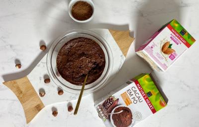 Foto em tons de branco da receita de creme de avelã caseiro servida em um bowl de vidro sobre uma tábua de madeira com embalagens de bebida vegetal de amêndoa e cacau em pó NATURE'S HEART