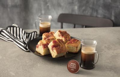 Fotografia em tons de marrom em uma bancada de madeira com um pano de prato com listra marrons, um prato redondo raso marrom com pedaços de bolo de goiabada. Ao lado, uma xícara de café Nescafé Dolce Gusto.