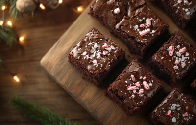 Fotografia vista de cima de vários quadrados de brownie com confeitos de natal, sobre uma tábua de madeira que está apoiada em uma mesa de madeira que tem luzes de natal.