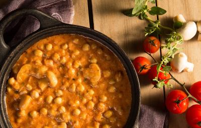 Fotografia em tons de amarelo em uma bancada de madeira com um pano azul e uma panela de ferro preta com alças laterais e o strogonoff de grão-de-bico dentro. Ao lado, tomates, champignon e salsinha espalhados na bancada.