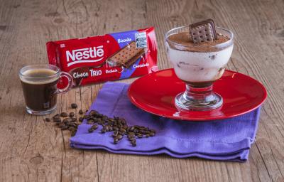 Fotografia de uma taça transparente sobre um prato redondo vermelho, preenchida com um creme branco, um pedaço de choco trio e cacau polvilhado por cima