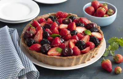 Fotografia em tons de vermelho e azul de uma bancada, ao centro a torta com morango, amoras e cerejas. Ao lado um pano quadriculado azul e branco e morangos.