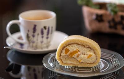 Fotografia de um rocambole feito de pão de ló de chocolate recheado com chantilly e peras cozidas, que está em um prato pequeno de vidro. Ao fundo, tem uma xícara de chá branca, sobre um miniprato de vidro branco, ao lado de uma colher de chá.