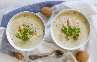 Fotografia vista de cima de dois recipientes na cor branca com um creme de aveias com palmito, pimenta por cima e salsinha. Ao redor, três biscoitos e uma colher de sopa, sobre uma toalha de mesa branca com faixas em azul.