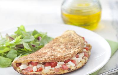 Fotografia em tons de verde em uma mesa de madeira cinza com um pano verde, um prato branco raso redondo e a omelete integral recheada com queijo branco, tomate e ervas, ao lado salada para acompanhar. Ao fundo, um potinho com azeite.