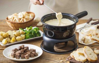 Fotografia em tons de marrom e beje de uma bancada de madeira com uma panela de fondue preta com fondue de queijo. Ao lado um prato redondo com carne, um prato retangular com batatas e brócolis, e uma cesta com pães.