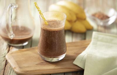 Fotografia em tons de marrom e amarelo de uma bancada de madeira com uma tábua, um paninho verde e um copo com a bebida dentro e um canudo de papel amarelo. Ao fundo uma jarra com a bebida, bananas e cacau.