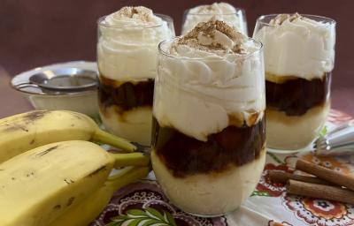 Foto da receita de banana cremosa servida em 4 porções em copos largos de vidro, sobre uma mesa decorada com uma toalha colorida, algumas bananas em cacho e paus de canela. Ao fundo um bowl de porcelana.