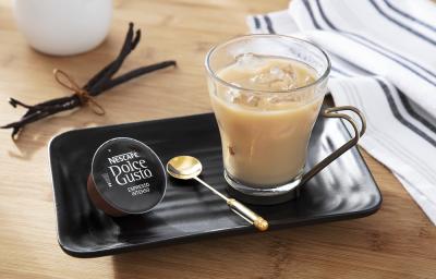 Foto em tons de bege da receita de espresso com baunilha e leite gelado servida em uma xícara de vidro sobre uma base de porcelana preta com uma colher dourada e uma cápsula de dolce gusto espresso intenso