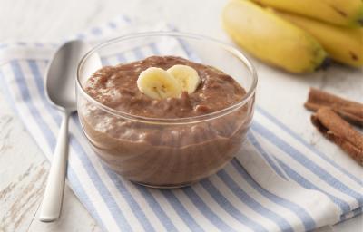 Fotografia em tons de azul em uma bancada de madeira clara com um paninho listrado azul e em cima um potinho de vidro com o doce de banana e cacau. Ao lado, algumas bananas, uma colher e canela em pau.