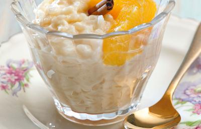 fotografia em tons de branco e amarelo tirada de um prato redondo branco com um copo transparente que contém arroz doce com rodelas de laranja de canela