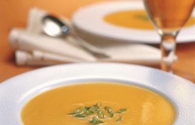 Fotografia em tons de laranja em uma mesa de madeira, com uma taça de vinho branco e um prato branco fundo com a sopa de abóbora, maçã e gengibre dentro dele.