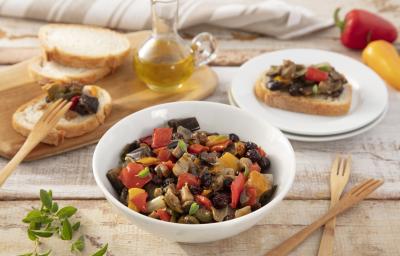 Fotografia em tons de vermelho e amarelo em uma bancada de madeira clara, um prato redondo branco grande ao centro com a caponata de berinjela. Ao lado, uma tábua de madeira com fatias de pães italianos, um potinho de azeite e pimentões amarelo e vermelho