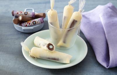 Foto da receita de geladinho algodão doce servida em 5 porções dentro de um pote transparente em cima de um prato de porcelana com um paninho lilás e algumas unidades de baton ao fundo