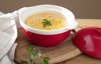 Fotografia de um recipiente pequeno e fundo, vermelho e branco, sobre uma tábua de madeira pequena ao lado da tampa e de uma pano branco. Dentro do recipiente tem o creme de cenoura e mandioquinha. Fotografia de um recipiente pequeno e fundo, vermelho e branco, sobre uma tábua de madeira pequena ao lado da tampa e de uma pano branco. Dentro do recipiente tem o creme de cenoura e mandioquinha.
